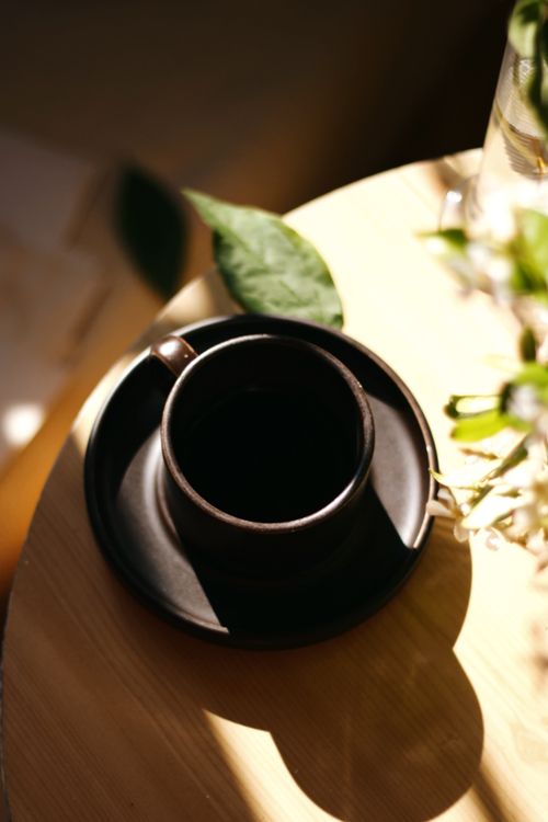 Free A warm coffee cup on a wooden table with sunlight streaming through leaves. Stock Photo