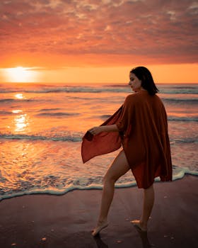Woman strolls along a beach at sunset wrapped in a vibrant orange cover, capturing summer's peaceful essence.