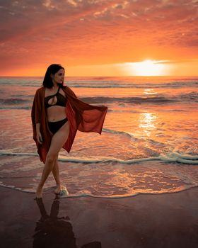 Woman in bikini on sunset beach, enjoying serene ocean ambiance with vibrant orange sky.