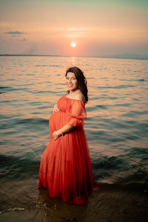 Free A Pregnant Woman in Red Dress Standing on Shore Stock Photo