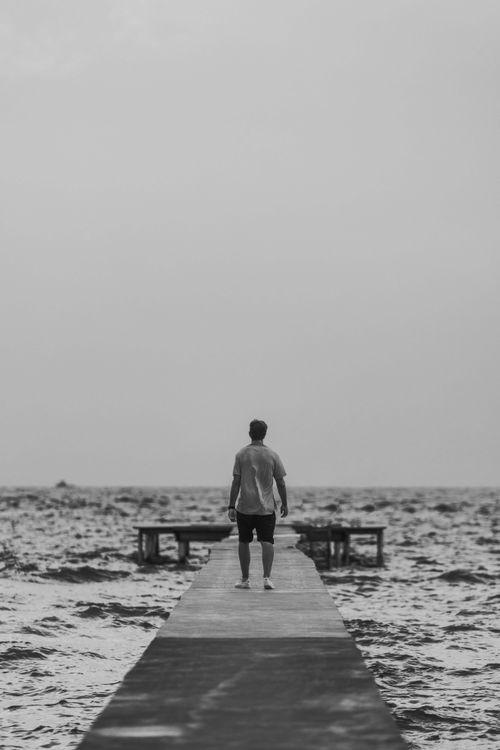 Free A monochrome image of a man standing alone on a dock over the sea, evoking solitude. Stock Photo