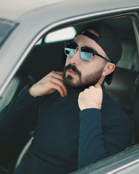 A fashionable man wearing sunglasses and a cap sits confidently inside a car.
