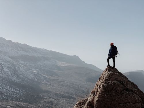 Free An adventurous solo hiker stands on a rocky peak, overlooking vast mountainous terrain at sunrise. Stock Photo