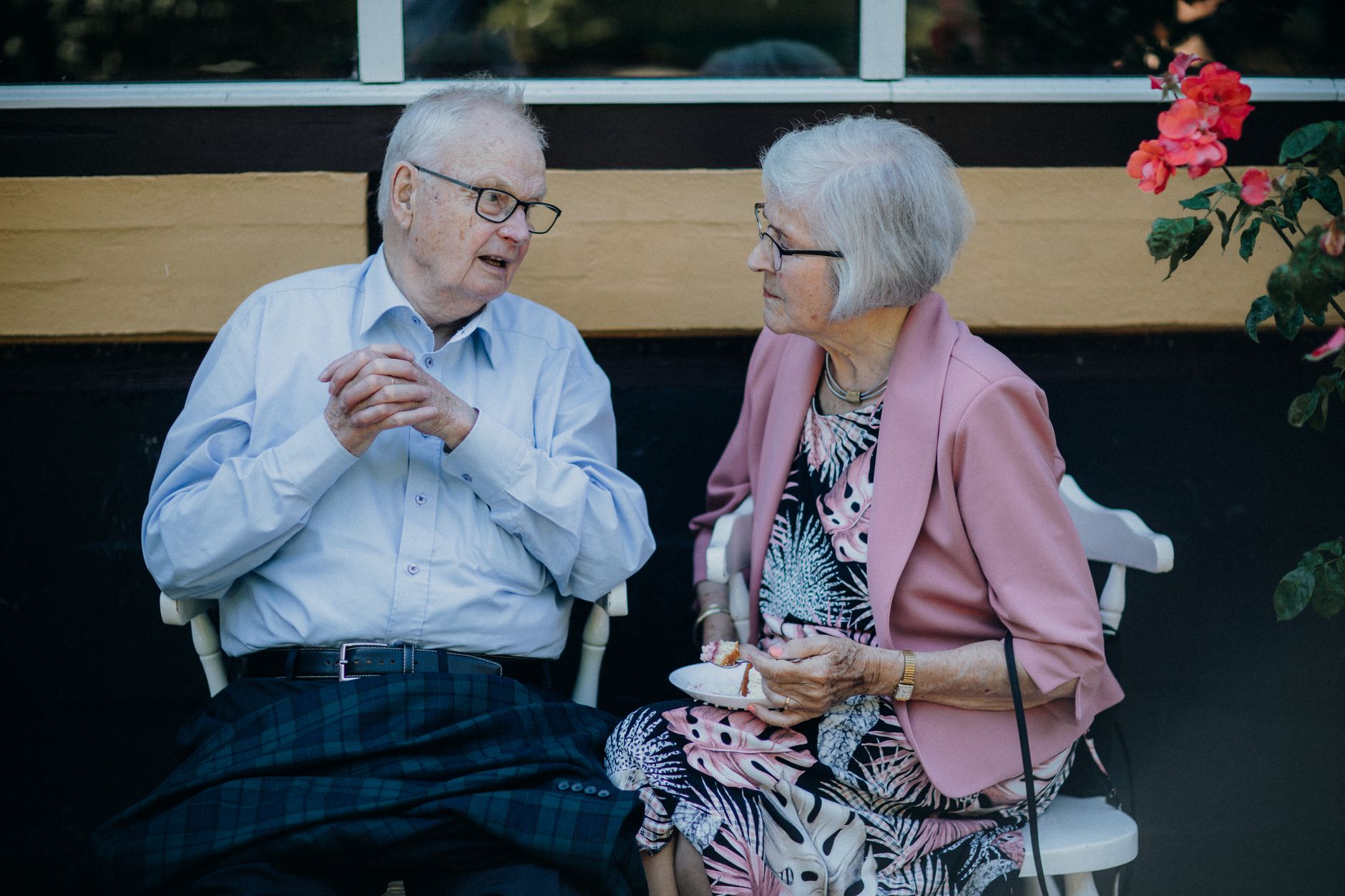 Senior couple sitting outside, engaged in conversation with flowers nearby. Captures a candid moment.
