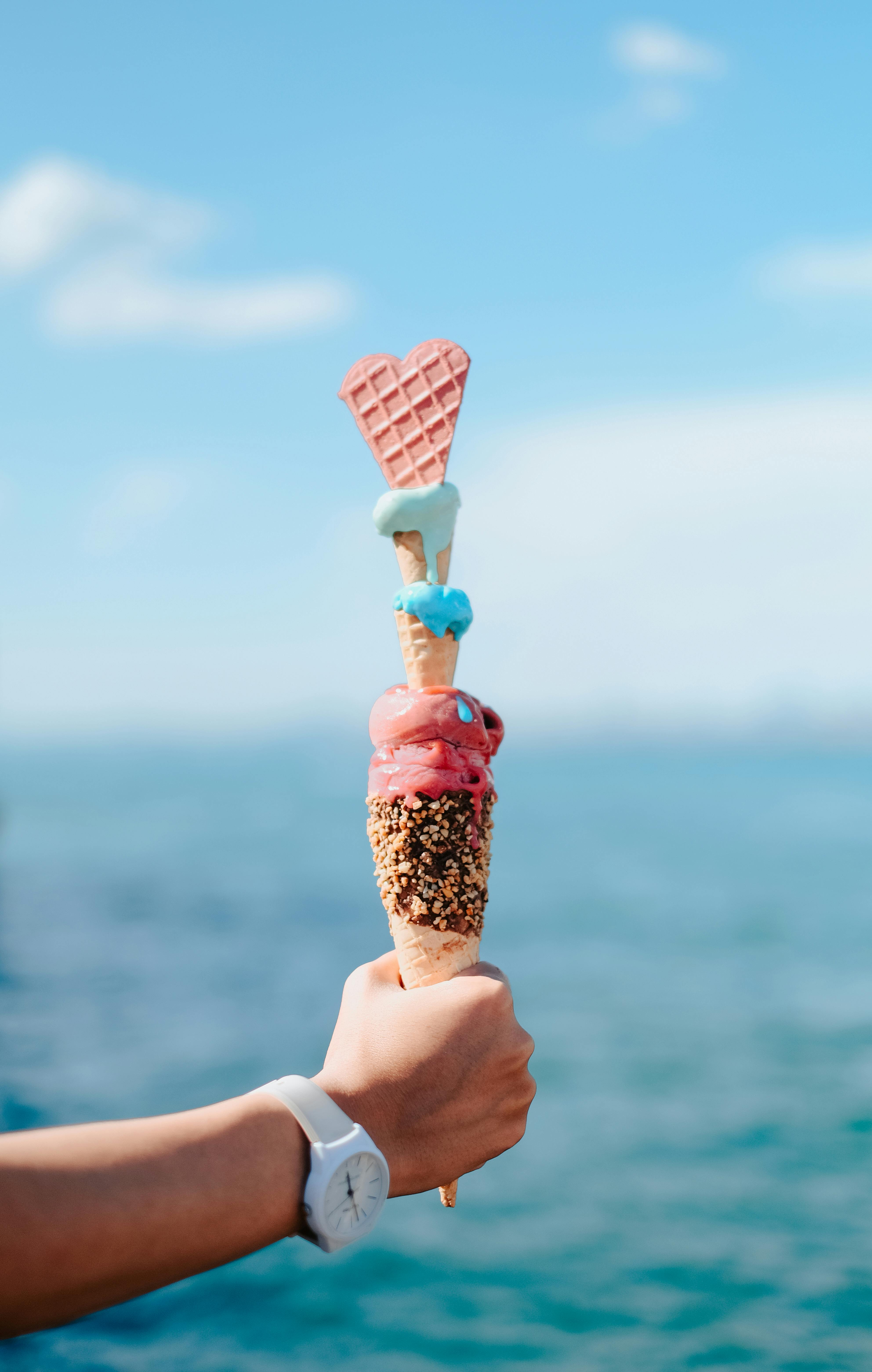 Triple-scoop ice cream cone with heart-shaped wafer held against the ocean.