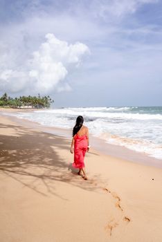 A woman in a red dress walks along a sunny beach with waves and palm trees.