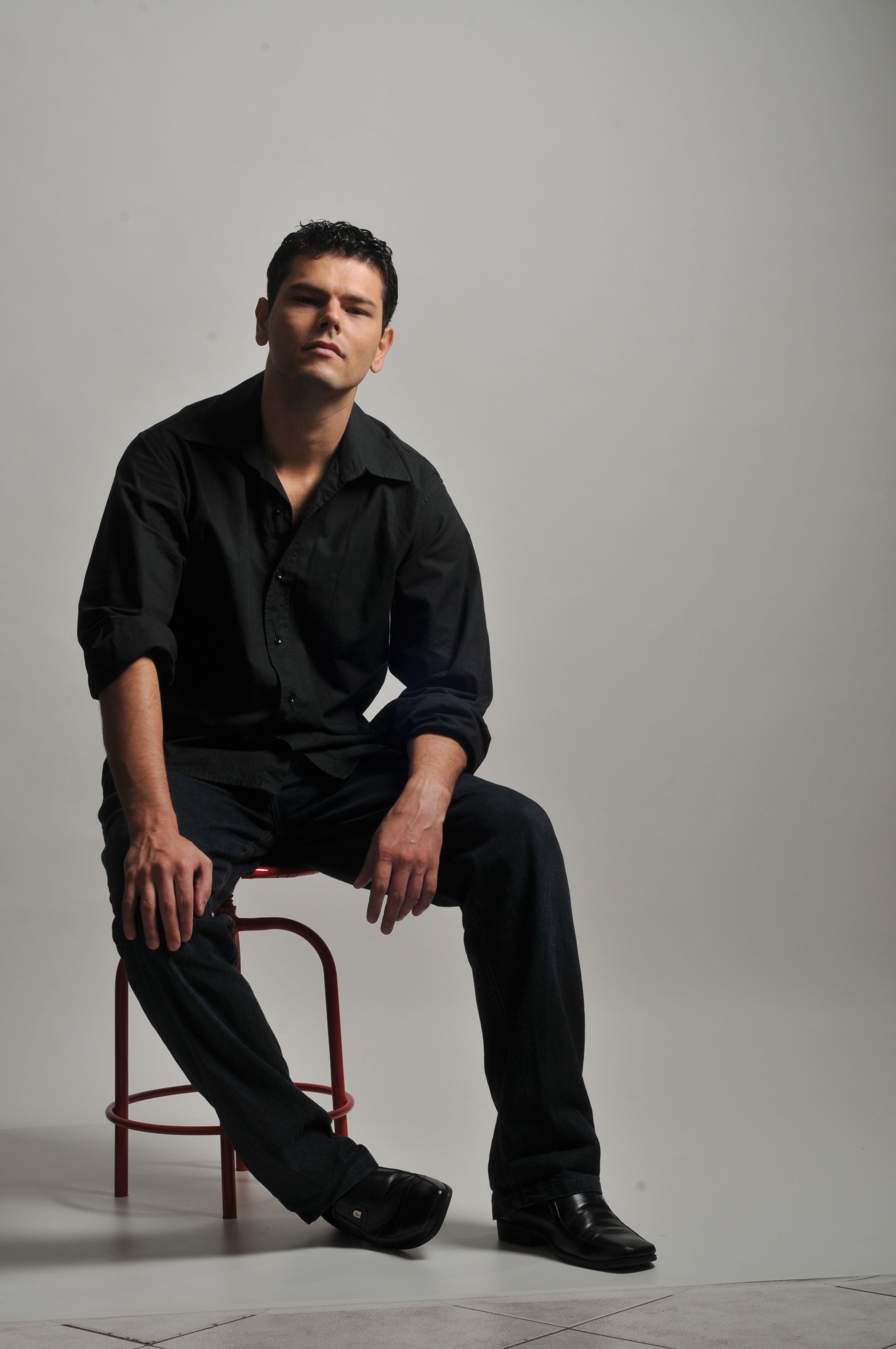 A man in black clothing poses stylishly on a red stool against a neutral background.
