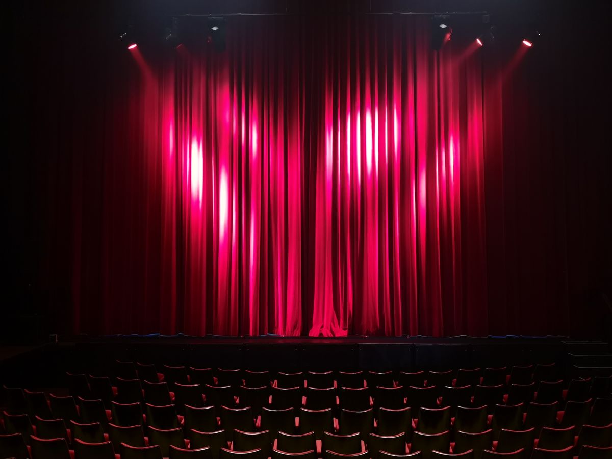 A closed red velvet theatre curtain lit by spotlights from above, with rows of empty dark seats in the foreground.