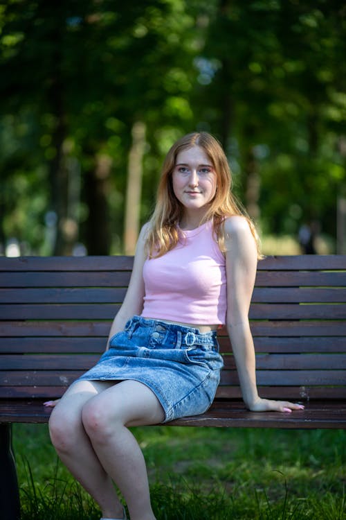 Woman in Pink Tank Top and Denim Skirt Sitting on Brown Wooden Bench