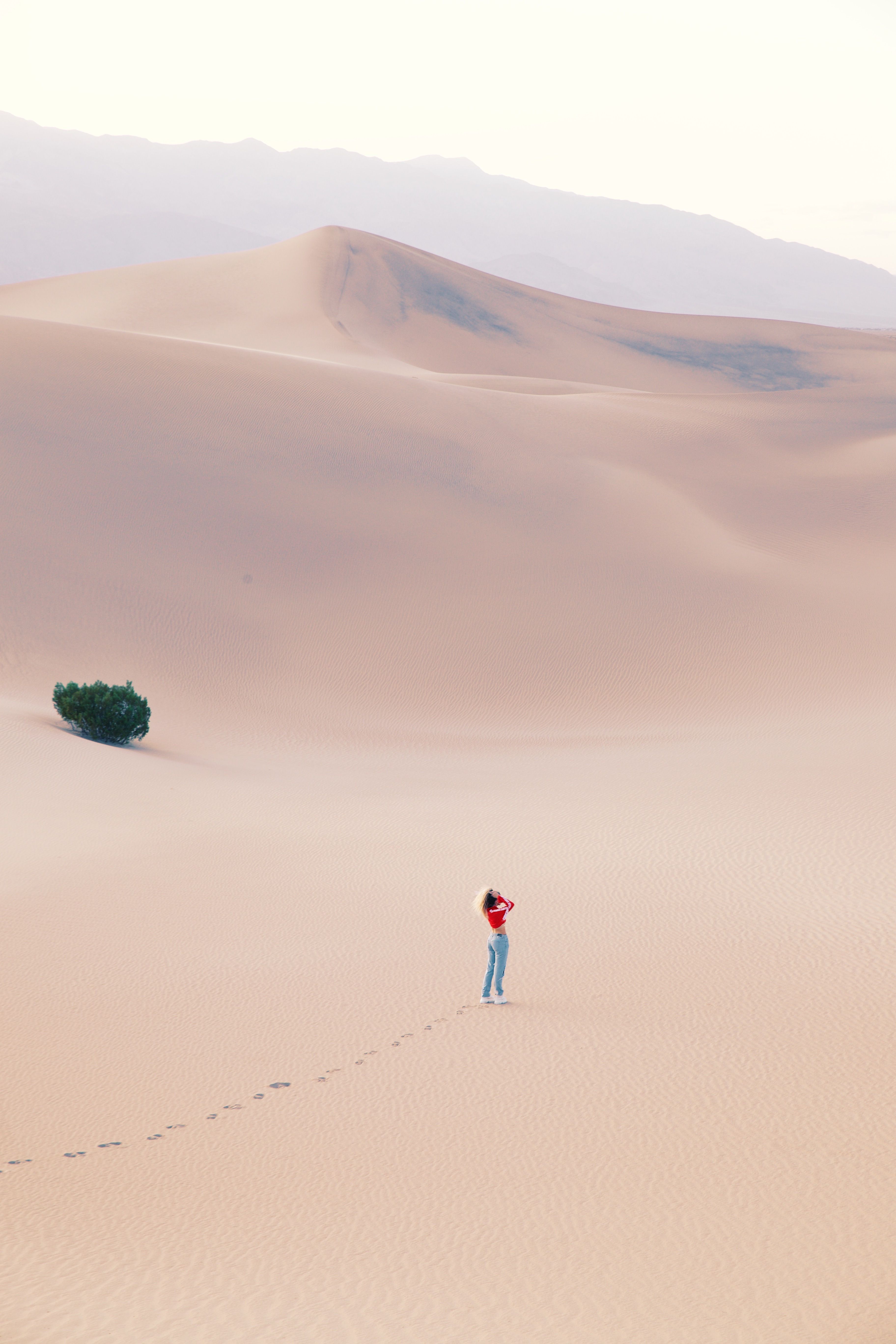 A lone woman stands in a vast desert, creating footprints in the sand under a pastel sky.