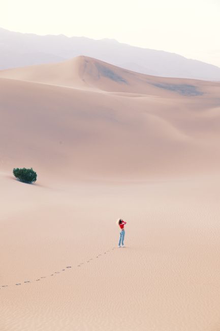 A lone woman stands in a vast desert, creating footprints in the sand under a pastel sky.