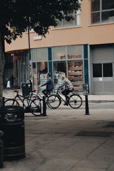 Two adults cycling past a London bookstore in a vibrant street setting.