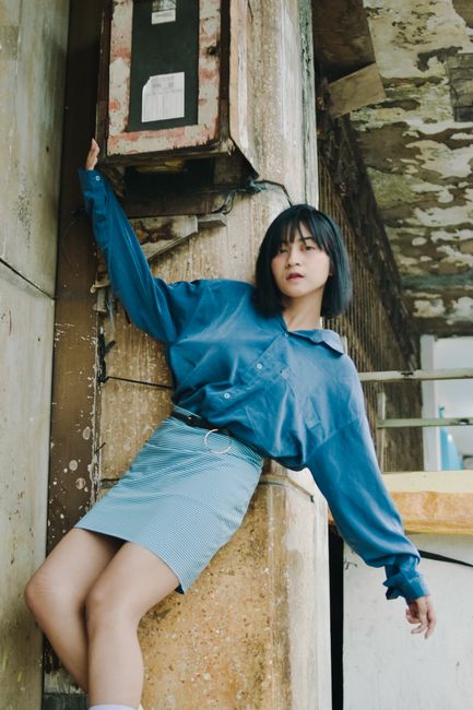 Young woman with bangs poses in a blue shirt and skirt against a rustic backdrop.