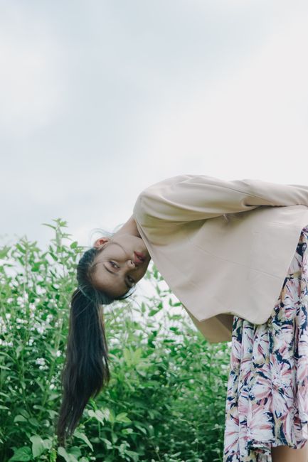 A young woman in a brown blazer and floral skirt bends playfully outdoors among green plants.