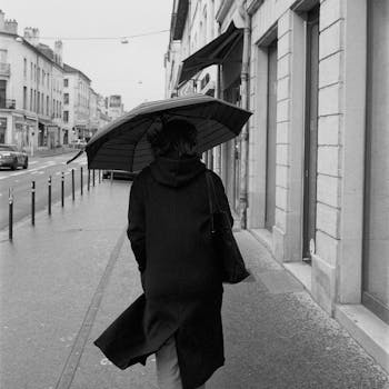 A person walks in the rain with an umbrella on a city sidewalk, captured in black and white.