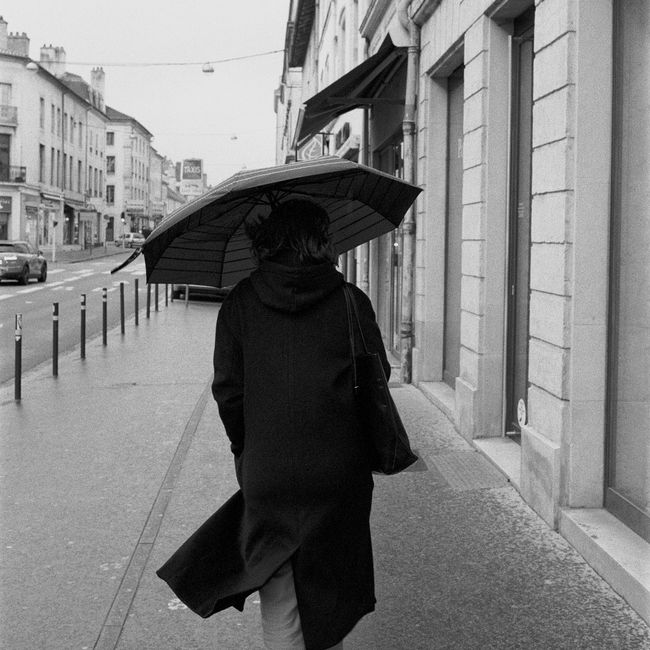 A person walks in the rain with an umbrella on a city sidewalk, captured in black and white.