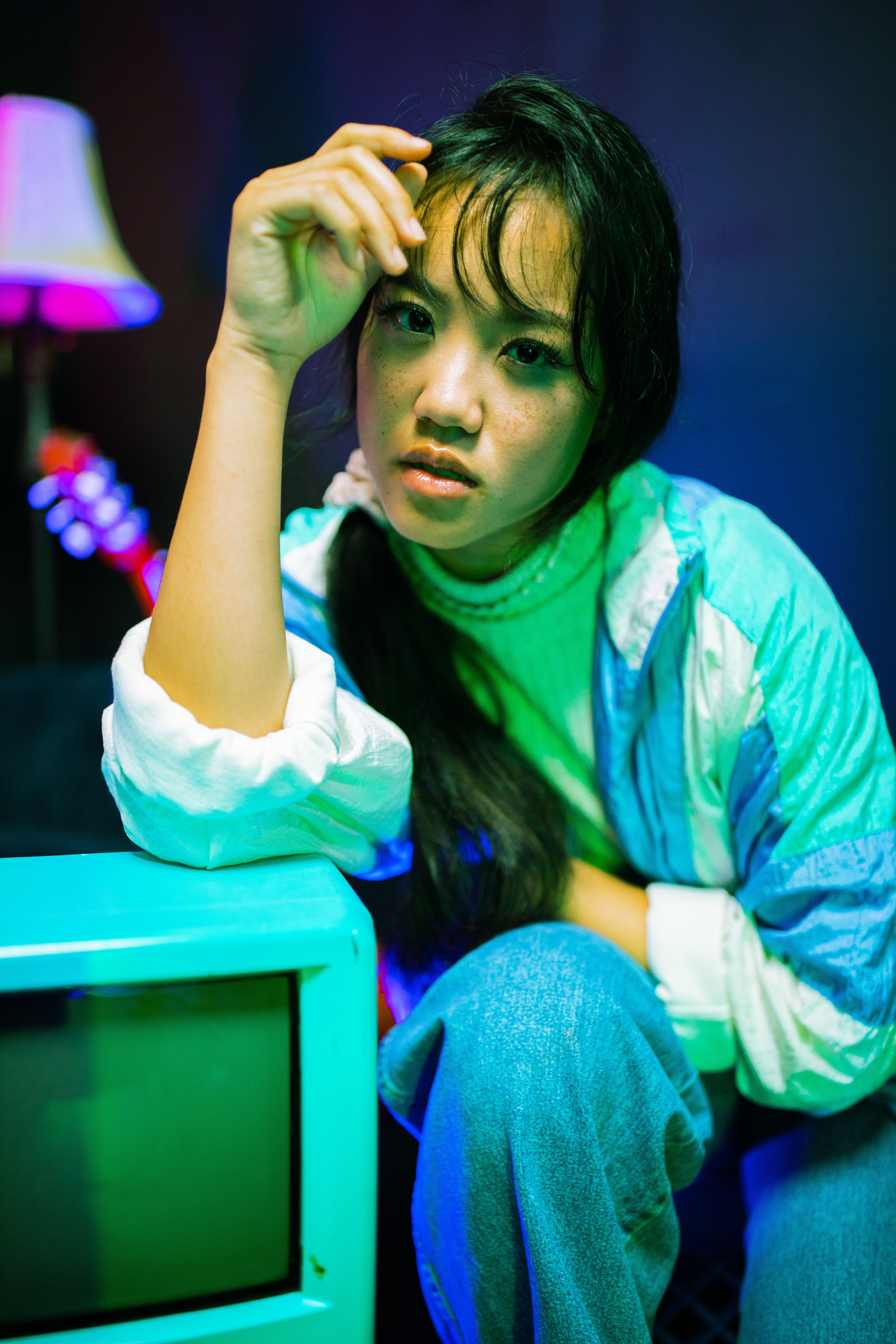 Asian woman with bangs poses by vintage TV in colorful lighting.