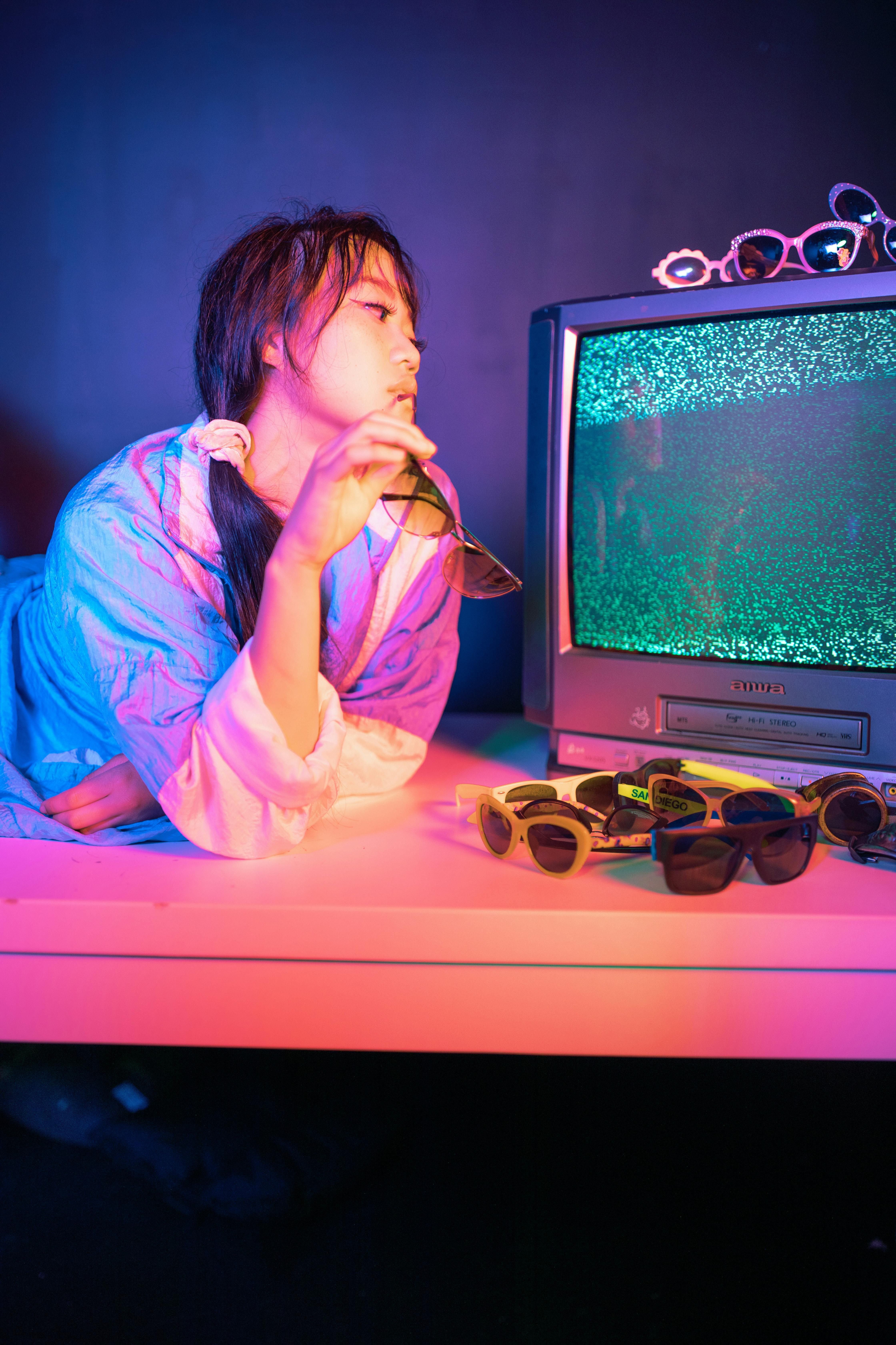 A woman poses in neon lights with vintage TV and sunglasses, capturing a retro vibe.