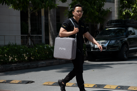 Asian man walking outdoors with a PS5 bag, showcasing urban lifestyle and fashion.