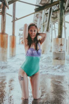 Woman in colorful swimsuit kneeling under a pier at the beach, surrounded by waves.