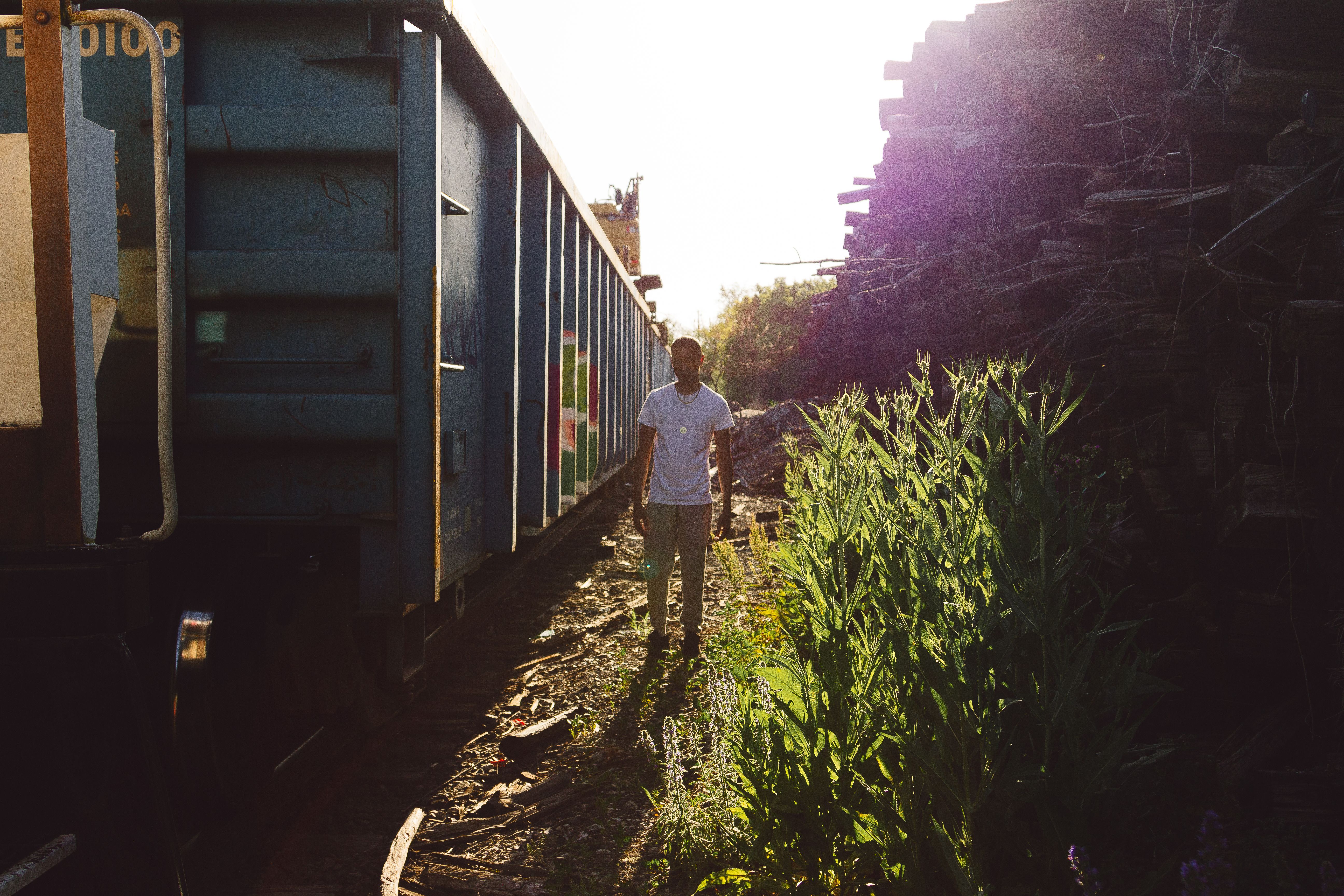 A man in casual attire walking beside a cargo train in the sunlight, surrounded by nature.