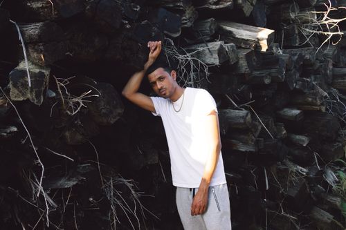 Free Portrait of a man in white t-shirt and grey pants leaning against stacked logs outdoors. Stock Photo