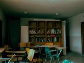 An empty classroom with scattered chairs and a bookshelf full of old books.