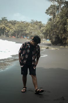 A man walks thoughtfully on a peaceful tropical beach, surrounded by lush green trees and crashing waves.