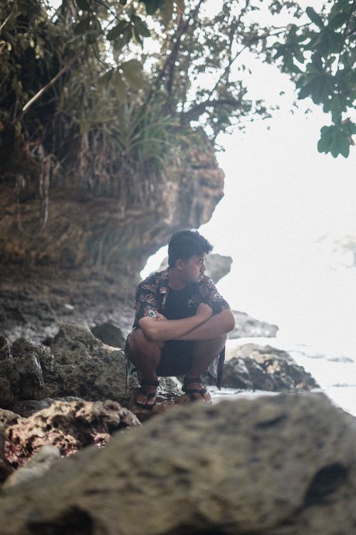 Free A man crouches on a rocky cliff near the sea, surrounded by lush foliage. Stock Photo