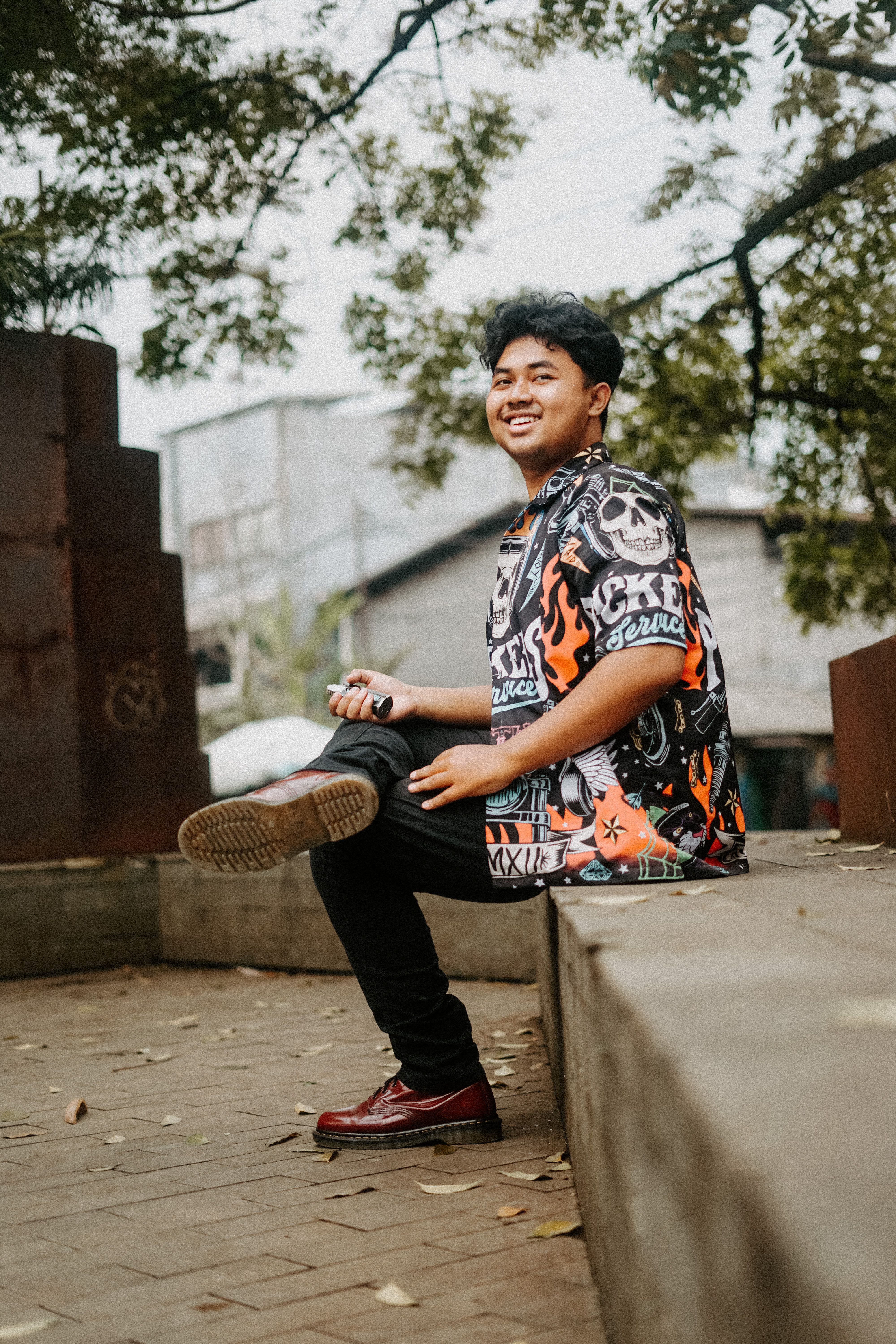 A cheerful young man wearing a bold shirt sits smiling in an urban park.