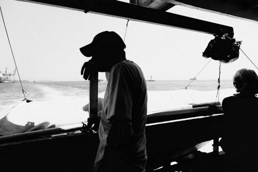 A silhouetted fisherman stands thoughtfully on a boat with industrial ports in the distance.