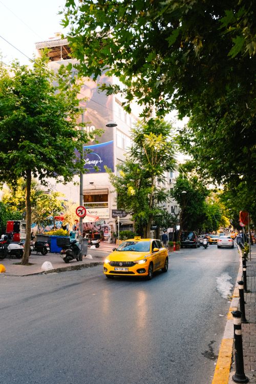 Free A lively urban street scene with a yellow taxi and lush green trees in the afternoon light. Stock Photo