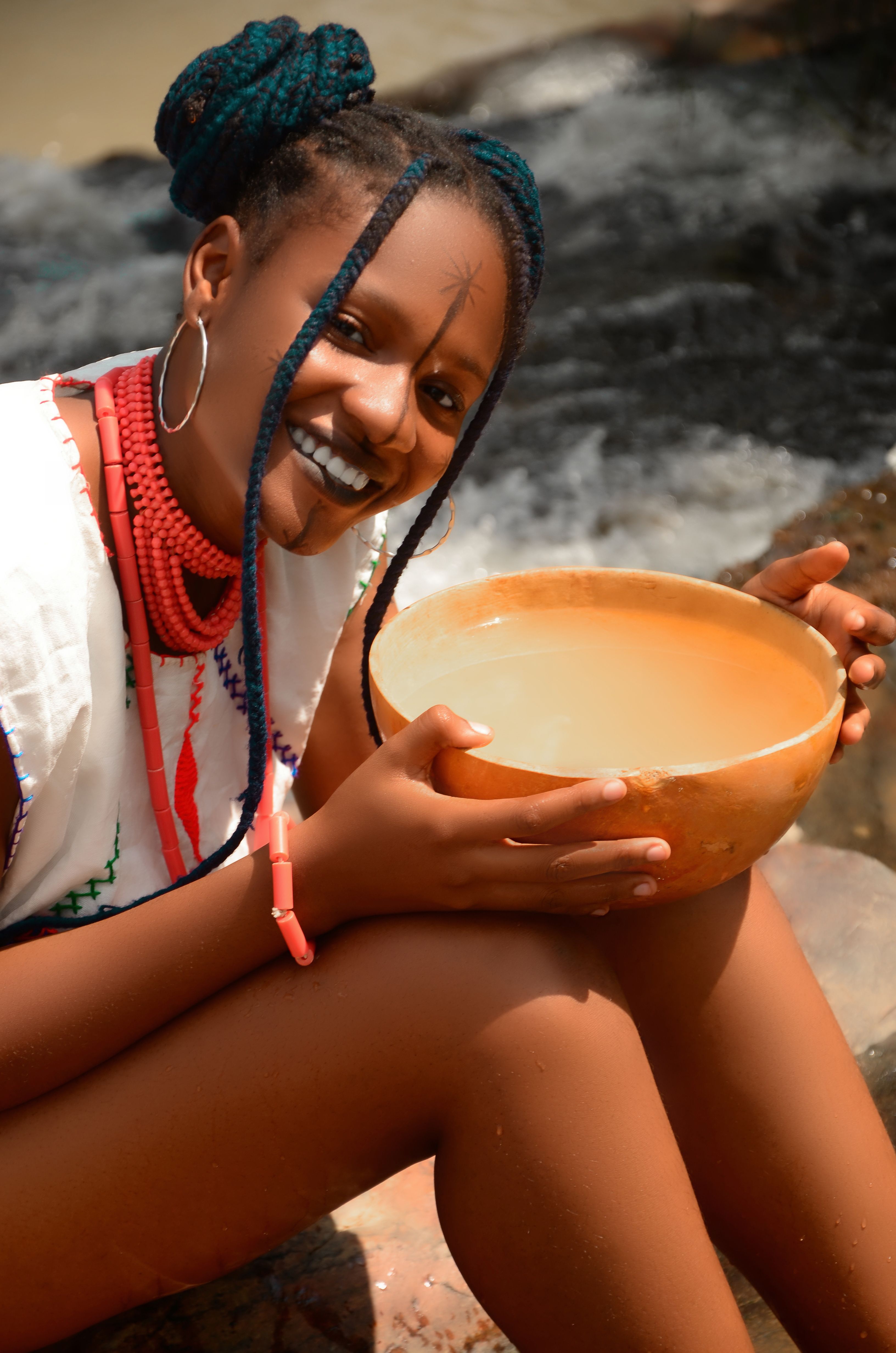 An African woman with braided hair holding a bowl, smiling near a waterfall.