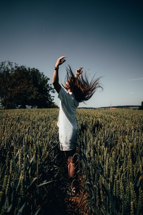 Free Woman in white dress stands with arms raised in a sunny wheat field, enjoying a bright summer day. Stock Photo