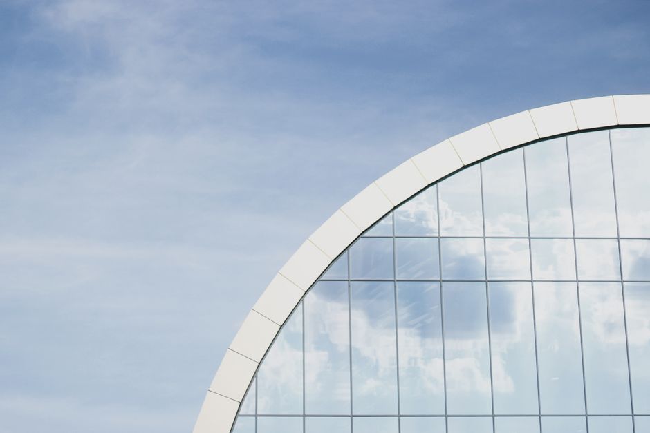 A modern glass building with reflective windows showing clouds against a blue sky.