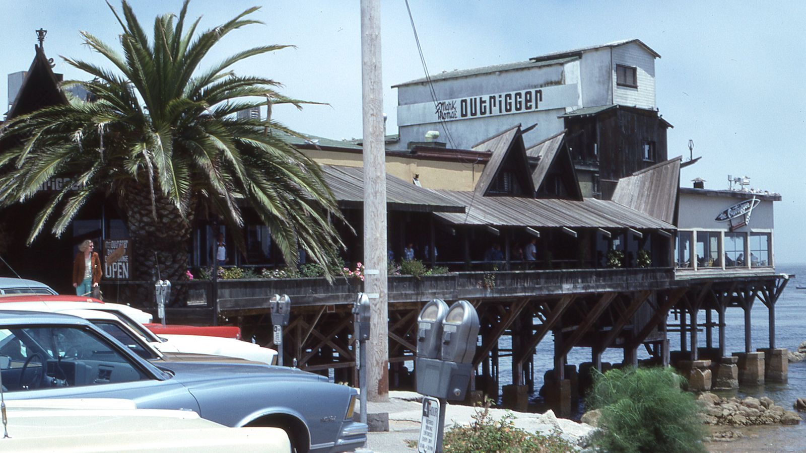 Retro-style coastal restaurant with palm tree and parked cars on a sunny day.