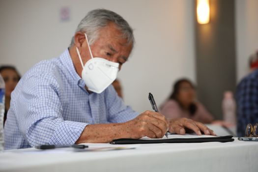 Elderly man with face mask sitting indoors, focused on writing in a notebook. - Fotografer Sidoarjo
