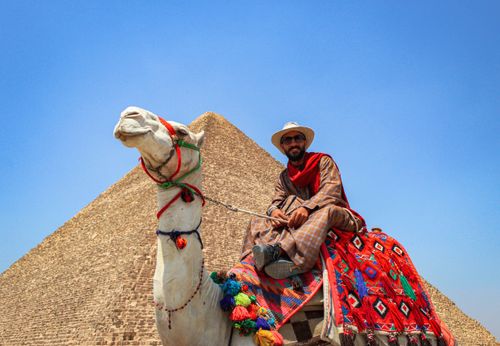 Free A man in traditional attire rides a camel near the Great Pyramid of Giza under a clear blue sky. Stock Photo