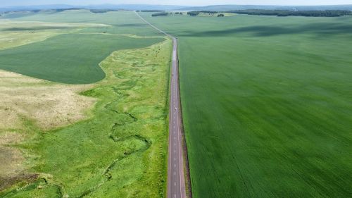 Free Drone captures a long road cutting through expansive green fields on a clear day. Stock Photo