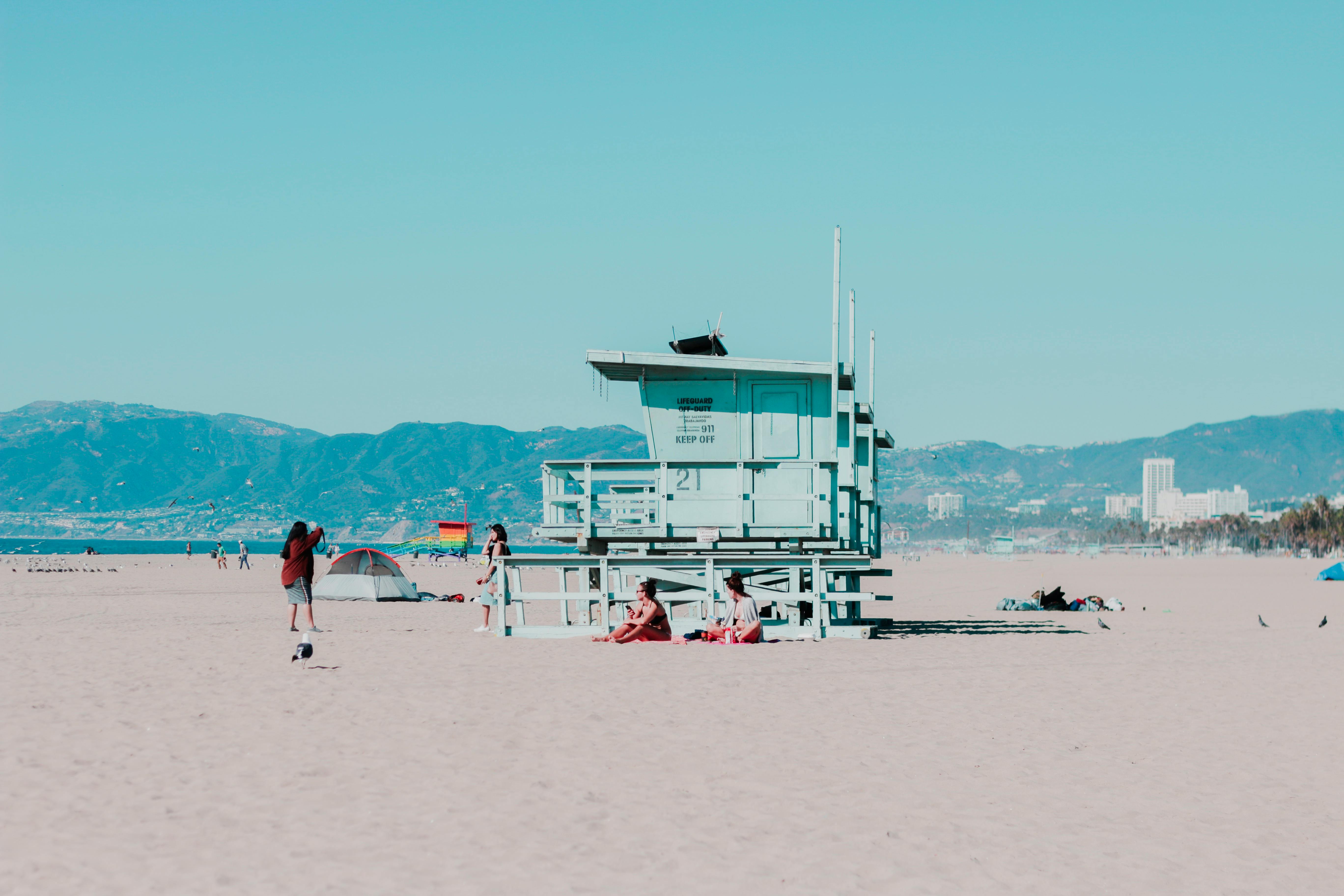 people sitting on sand near white wooden cottage near seashore