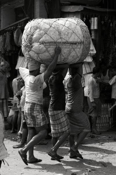 Three men carrying a heavy bundle on their shoulders through a market