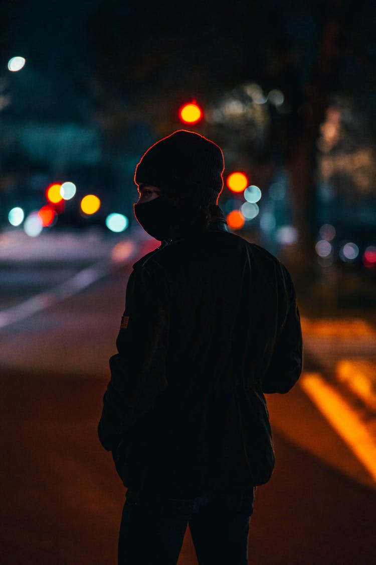 A Person Wearing Beanie And Face Mask Standing On The Street At Night