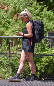 Side view of a woman with a backpack checking her phone while hiking in a lush, sunny setting.
