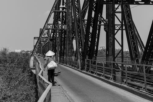 A Grayscale Photo of a Person in Plaid Jacket Walking on the Bridge