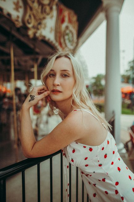 Portrait of a young woman in a cherry-patterned dress posing by a carousel at an amusement park.