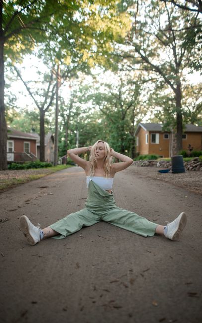 Woman in green overalls and sneakers sitting on a road, surrounded by trees and houses.