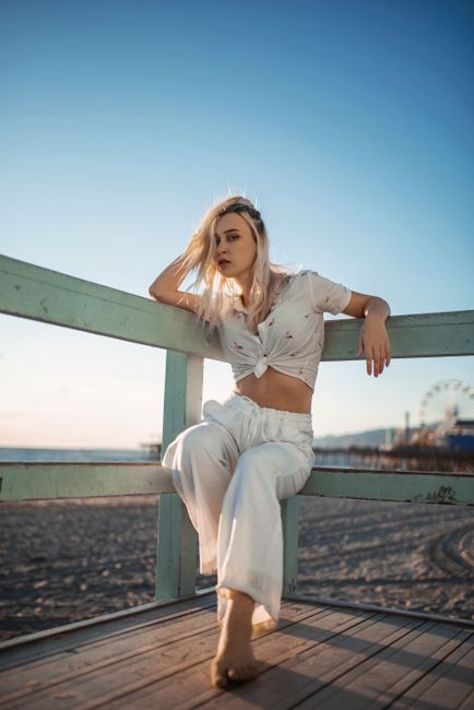 Blonde woman posing at a beach pier, wearing white summer outfit, during sunset.