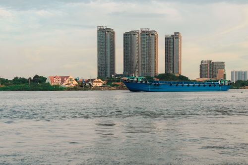 Free A serene view of a blue cargo ship passing city buildings on a calm river at sunset. Stock Photo