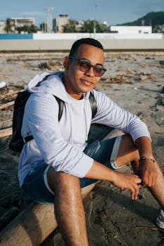 A man in sunglasses sits on a sandy beach wearing a hoodie and shorts, enjoying the sunny day.