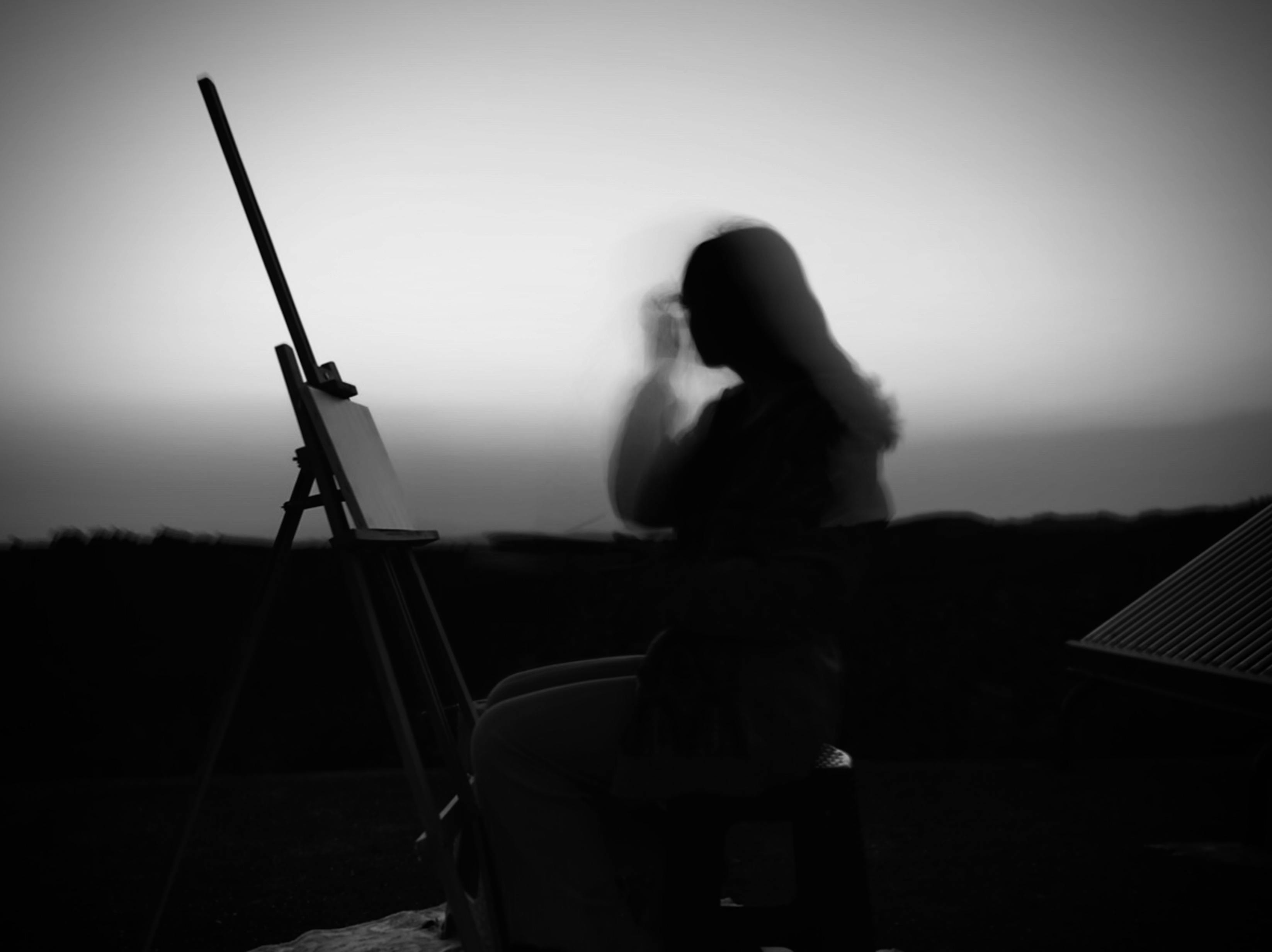 A blurred silhouette of a woman painting on an easel at dusk, creating a mysterious mood.